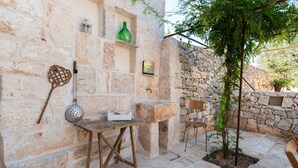 A stone-walled outdoor area with a wooden table, a hanging basket, and a small tree. - Masseria Fumarola Il Cavaliere IV by Rentbeat (Ostuni)