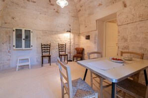 A rustic stone-walled room with a dining table, chairs, a small cabinet, and a light fixture. - Masseria Fumarola Il Cavaliere IV by Rentbeat (Ostuni)