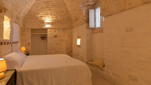 A stone-walled bedroom with a bed, bedside lamps, and a small window. - Masseria Fumarola Il Cavaliere IV by Rentbeat (Ostuni)