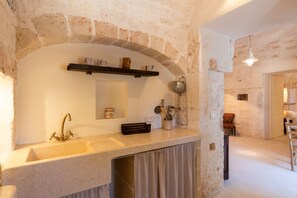 A stone-walled kitchen with a sink, a mirror, and a shelf with kitchen items. - Masseria Fumarola Il Cavaliere IV by Rentbeat (Ostuni)