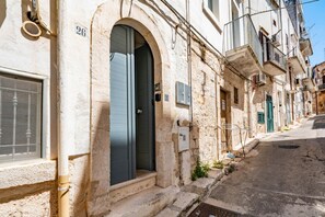 Una calle estrecha con un edificio que tiene un gran arco de entrada, una ventana con celosía metálica y un pequeño balcón con barandilla de metal.