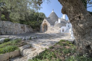 A stone building with a conical roof, a stone wall, and a tree in the foreground. - Trulli Capo di Lupo by Rentbeat (Cisternino)