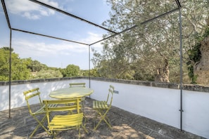 Un patio al aire libre techado con una mesa y sillas, rodeado por un muro blanco y árboles.
