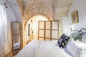 A bedroom with a stone archway, a bed with white linens, a wooden wardrobe, and a nightstand with a lamp.