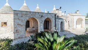 A stone building with multiple arches and conical roofs, a patio area with tables and chairs, and a garden with palm plants. - Trulli Garden Harmony by Rentbeat (Martina Franca)