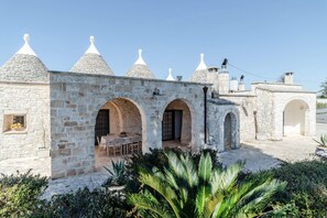 A stone building with multiple arches and conical roofs, a patio area with tables and chairs, and a garden with palm plants.