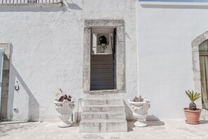 A white building with a small window, a set of stairs leading to the entrance, and two potted plants.