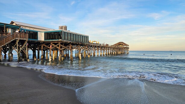 A pier extending into the ocean with a building on it, a beach in the foreground, and a clear sky.