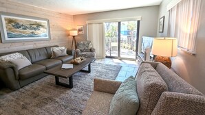 A living room with a sofa, armchair, and a coffee table. There is a painting on the wall, a lamp, and a view through the glass door.