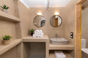 A bathroom with a stone sink, two round mirrors, a towel rack with rolled towels, and potted plants.