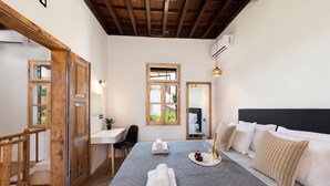 A bedroom with a bed, bedside table, chair, and a desk. There are windows, a mirror, and an air conditioning unit.