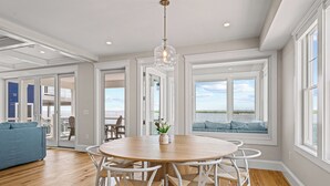 Un comedor amplio con una mesa redonda de madera y sillas blancas, ventanas grandes con vistas al océano y un sofá azul al fondo.