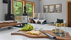 Una mesa de madera con una botella de vino Casillero del Diablo, uvas, queso y copas, ubicada en una habitación con ventanas amplias y una chimenea.