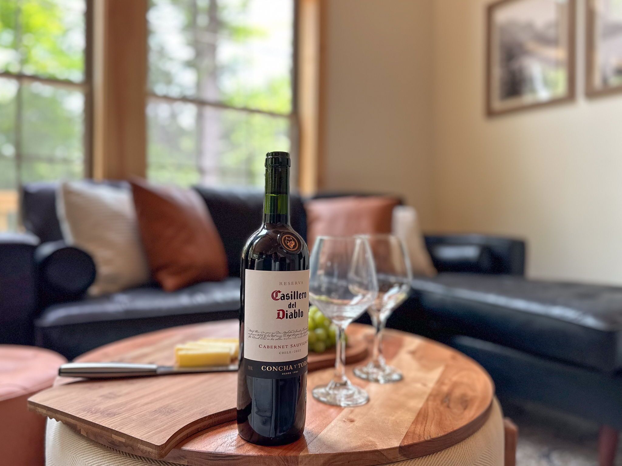 A bottle of Casillero del Diablo wine on a wooden table with a glass of wine and a knife, in a living room with a dark-colored sofa and a window with a view of trees.