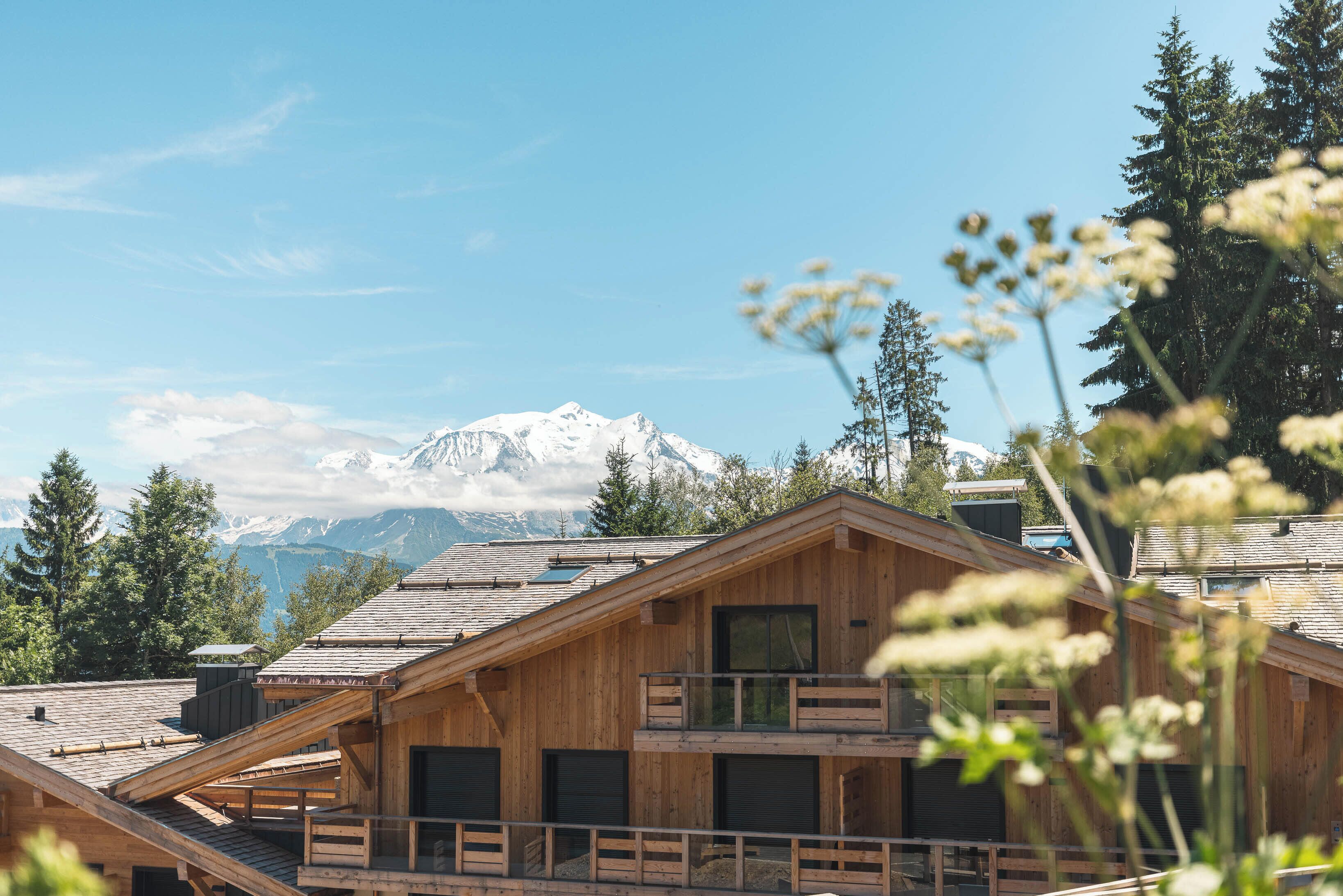A wooden cabin with a balcony, set against a backdrop of snow-capped mountains and a clear blue sky.
