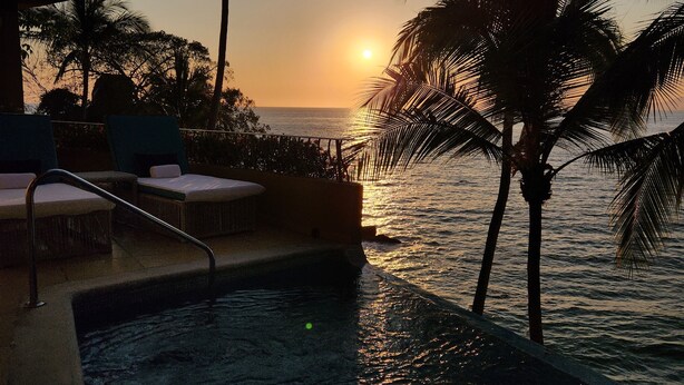 Blick auf ein Schwimmbecken mit Sonnenuntergang über dem Meer, Palmen und Liegestühlen.