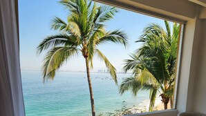 Ein Hotelzimmer mit großem Fenster mit Blick auf einen Strand und Palmen, ein Bett mit blauer Decke und ein Korbsessel.