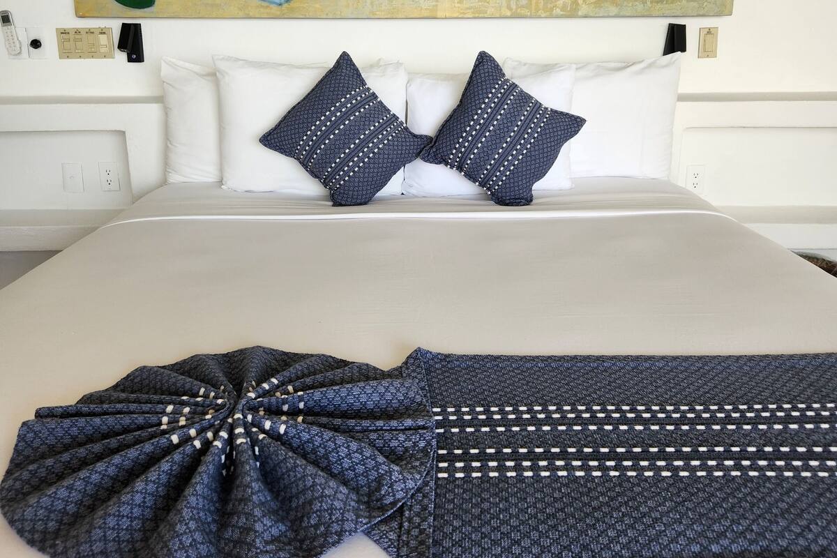 Une chambre d’hôtel avec un grand lit, du linge de lit blanc, deux coussins à motifs, un plaid bleu et blanc à motifs, et un tableau coloré au-dessus du lit.
