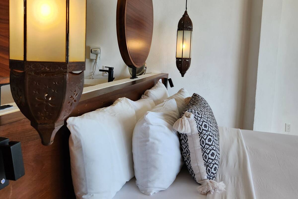 Une chambre d’hôtel avec un lit, des oreillers blancs, une tête de lit en bois, un miroir et deux lanternes fixées au mur.