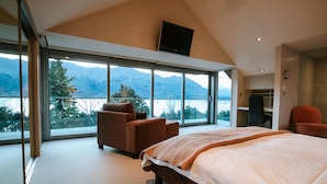 Ein Schlafzimmer mit einem großen Bett, einem Sessel, einem Schreibtisch und einem Fernseher. Es bietet Blick auf Berge und einen See.