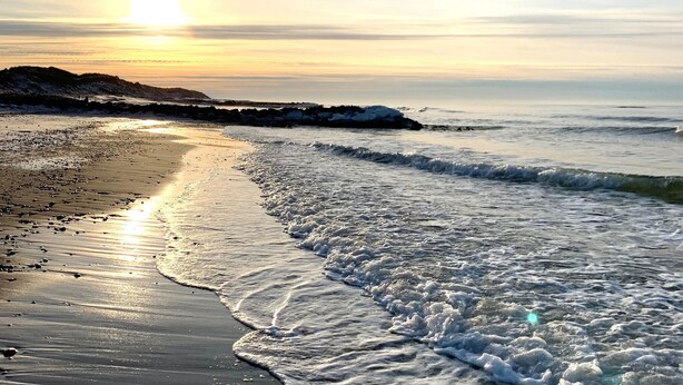 Une plage où les vagues viennent doucement lécher le rivage, un ciel dégagé et le soleil se couche ou se lève.