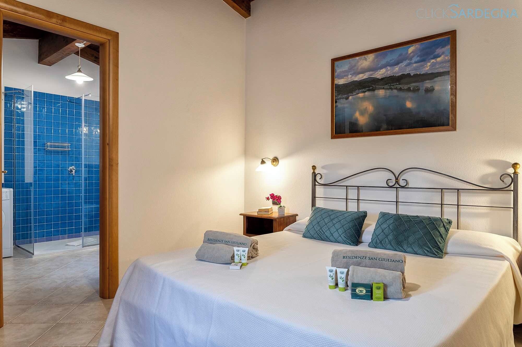 A hotel room with a bed, a nightstand, a painting, and a bathroom with blue tiles.