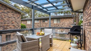 Ein überdachter Patio-Bereich mit Korbmöbeln, einem Grill und Blick auf Backsteingebäude.