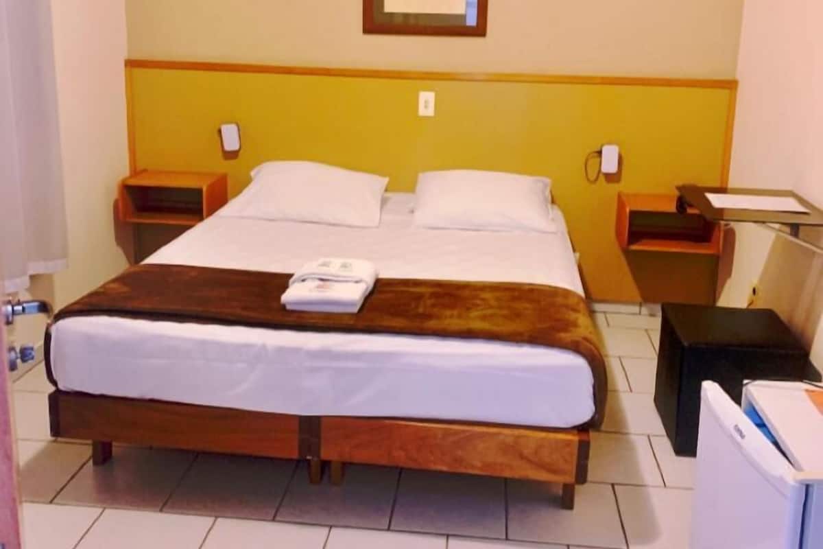 A hotel room with a double bed, two bedside tables, a nightstand, a framed picture on the wall, and a small desk with a chair.