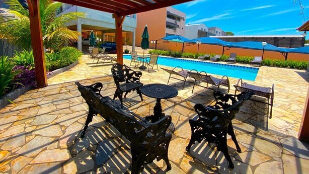 An outdoor patio with stone flooring, metal chairs, a round table, and umbrellas providing shade. A swimming pool is visible in the background. - Hotel Imigrantes LTDA (São Pedro)
