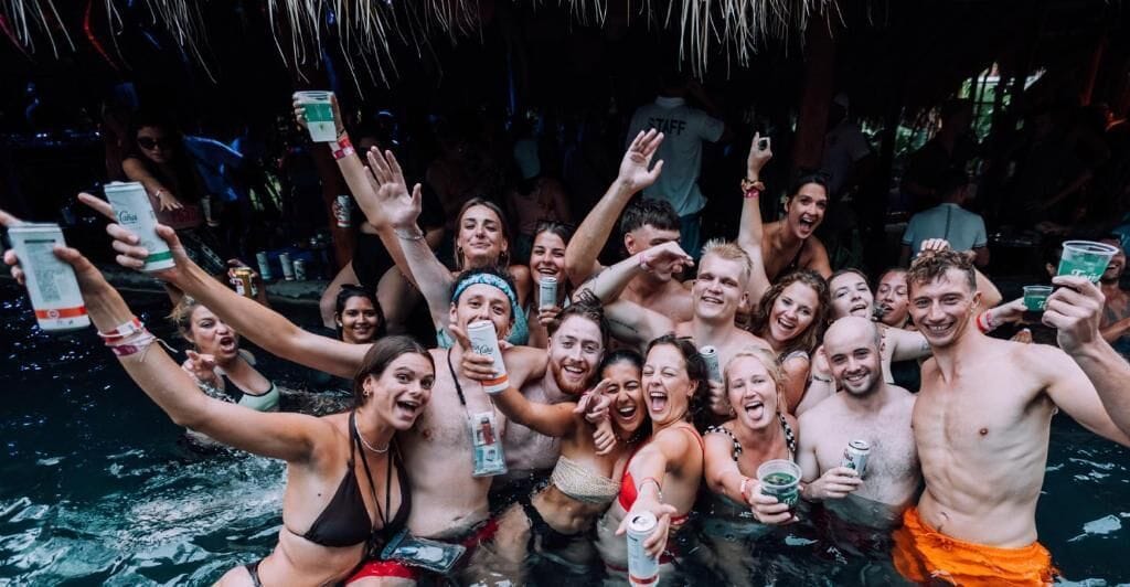 A group of people in a pool with drinks, some wearing swimwear, under a thatched roof.