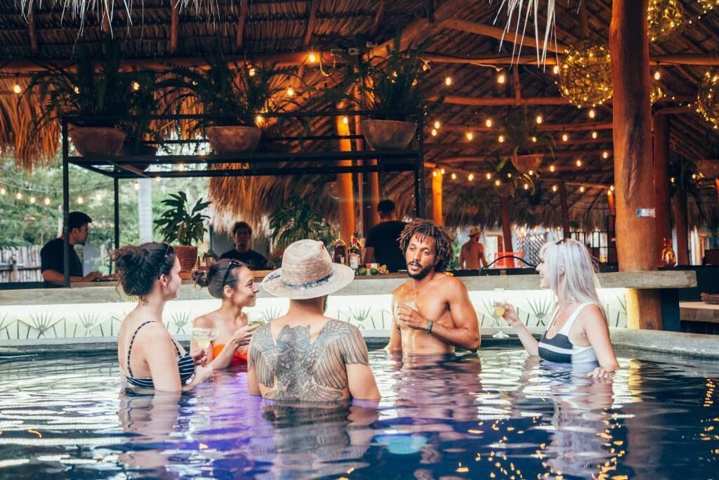 Un espace piscine où les gens se rencontrent et discutent, entouré d’une structure en bois agrémentée de guirlandes lumineuses et de plantes en pot.