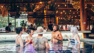 Un espace piscine où les gens se rencontrent et discutent, entouré d’une structure en bois agrémentée de guirlandes lumineuses et de plantes en pot.