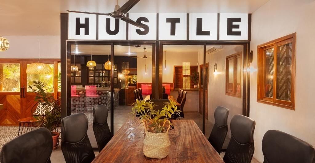 A conference room with a wooden table and black chairs, a 'HUSTLE' sign above the door, and a plant in the center.
