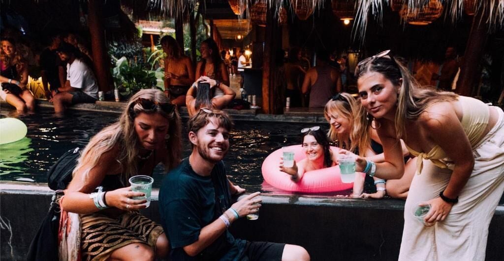 A group of people sitting by a pool with a tropical setting, including plants and a thatched roof structure.