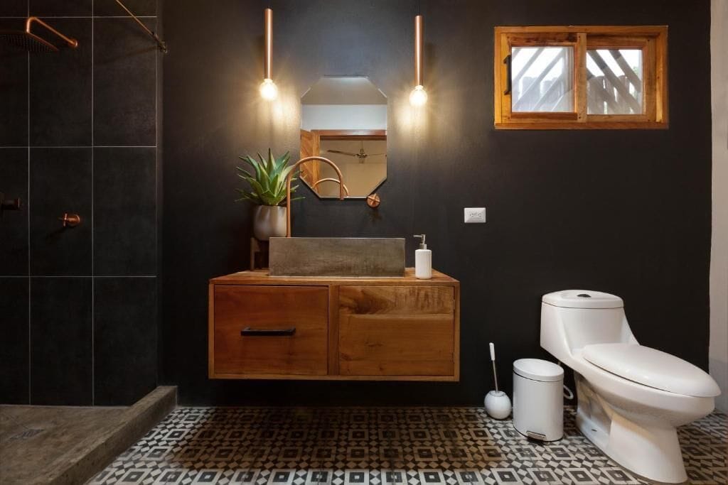 A modern bathroom with a dark tiled floor, a wooden vanity, a white toilet, and a small window.