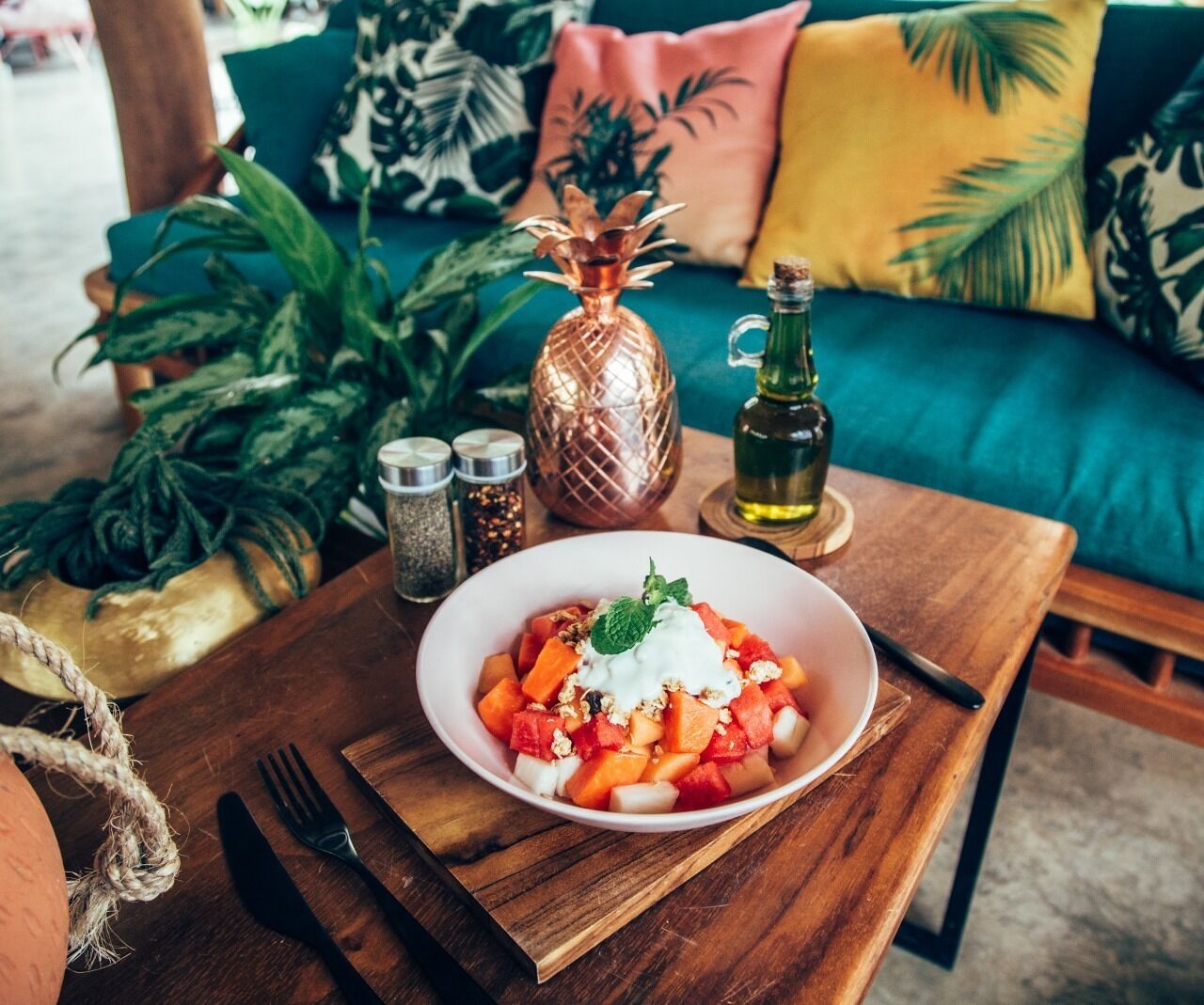 A wooden table with a bowl of fruit salad, a pineapple-shaped bottle, and a bottle of olive oil.