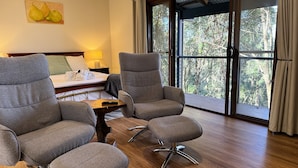 Une pièce comprenant deux fauteuils gris, une table en bois, un lit avec une tête de lit blanche, une table de chevet avec une lampe et une grande fenêtre donnant sur des arbres.