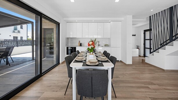 A modern kitchen with white cabinets, a dining area with a long table set for six, and a staircase with a geometric railing. - Stunning home in Vodice with WiFi (Vodice)