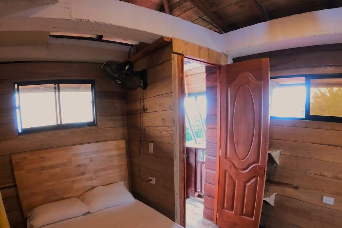 A wooden cabin room with a bed, a small window, and a door leading outside.