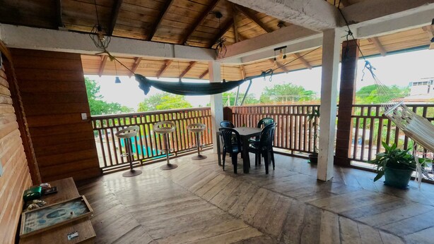 A covered outdoor area with a wooden floor, a hammock, a table with chairs, and a railing. - Petra Hotel (Nuquí)