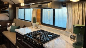 Et kompakt campingvognkøkken med kogeplade, lille spiseplads og et vindue med gardiner.