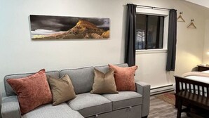 A modern living room with a grey sectional sofa, a wooden coffee table, and a large painting of a mountain landscape on the wall.