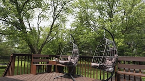 Une terrasse en bois avec deux fauteuils suspendus et une table, le tout entouré d’arbres.