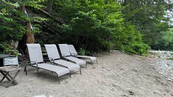 Une rangée de fauteuils de plage gris et une petite table sont disposées sur une plage de sable, entourées d’un feuillage verdoyant et dense.