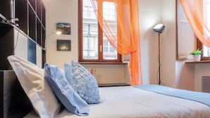 Une chambre à coucher avec un lit, des oreillers, une table de chevet, une lampe, une fenêtre avec des rideaux et un mur décoré de tableaux.