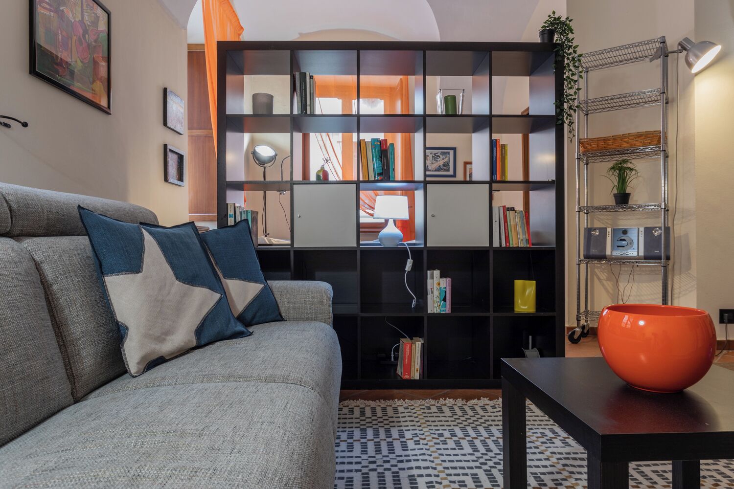 Un salon avec un canapé gris, une bibliothèque noire, une petite table sur laquelle se trouve un bol rouge, et un tapis à motifs.