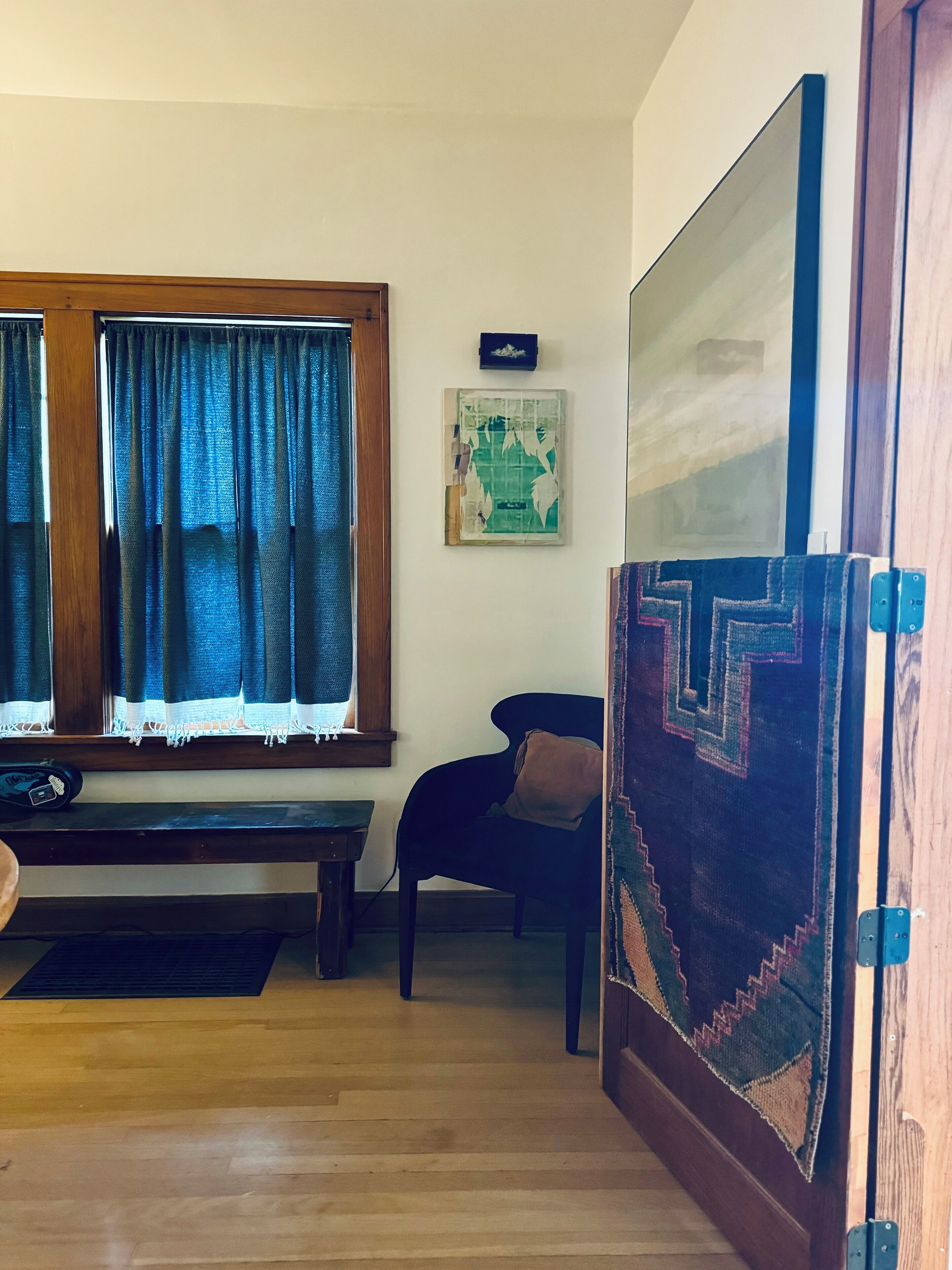 A room with a wooden floor, a bench, a chair, a window with curtains, a wall-mounted picture, and a door with a geometric-patterned rug.