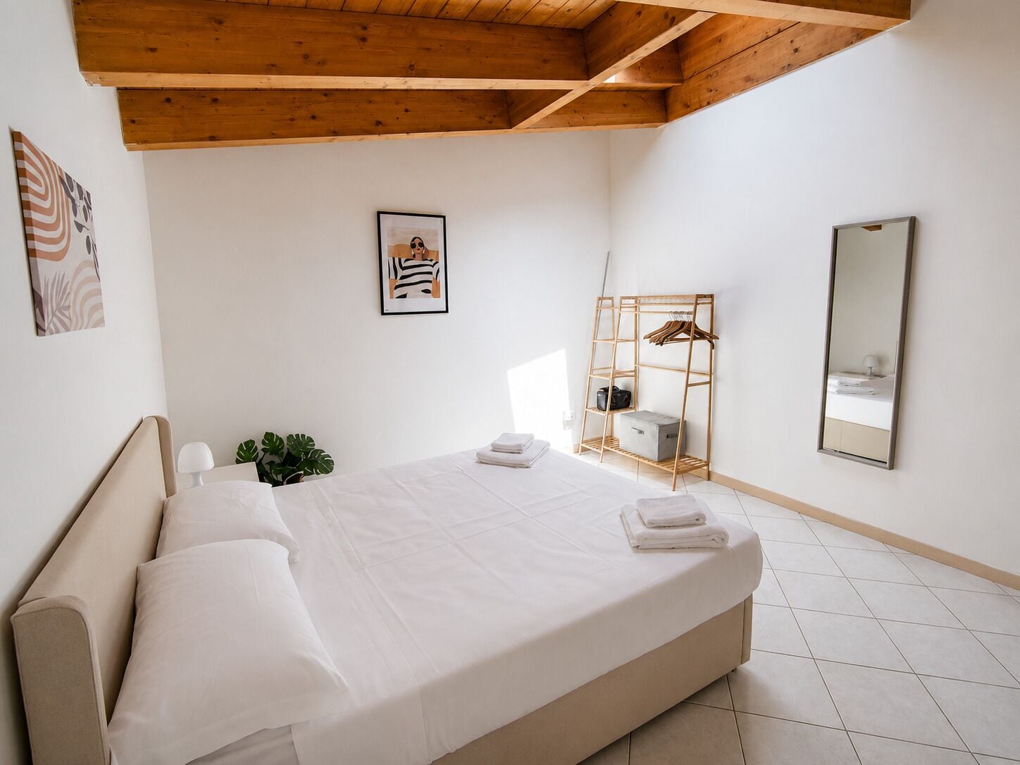 A hotel room with a bed, a wooden headboard, a wall-mounted mirror, a framed picture, and a wooden shelf with a hanging rack.