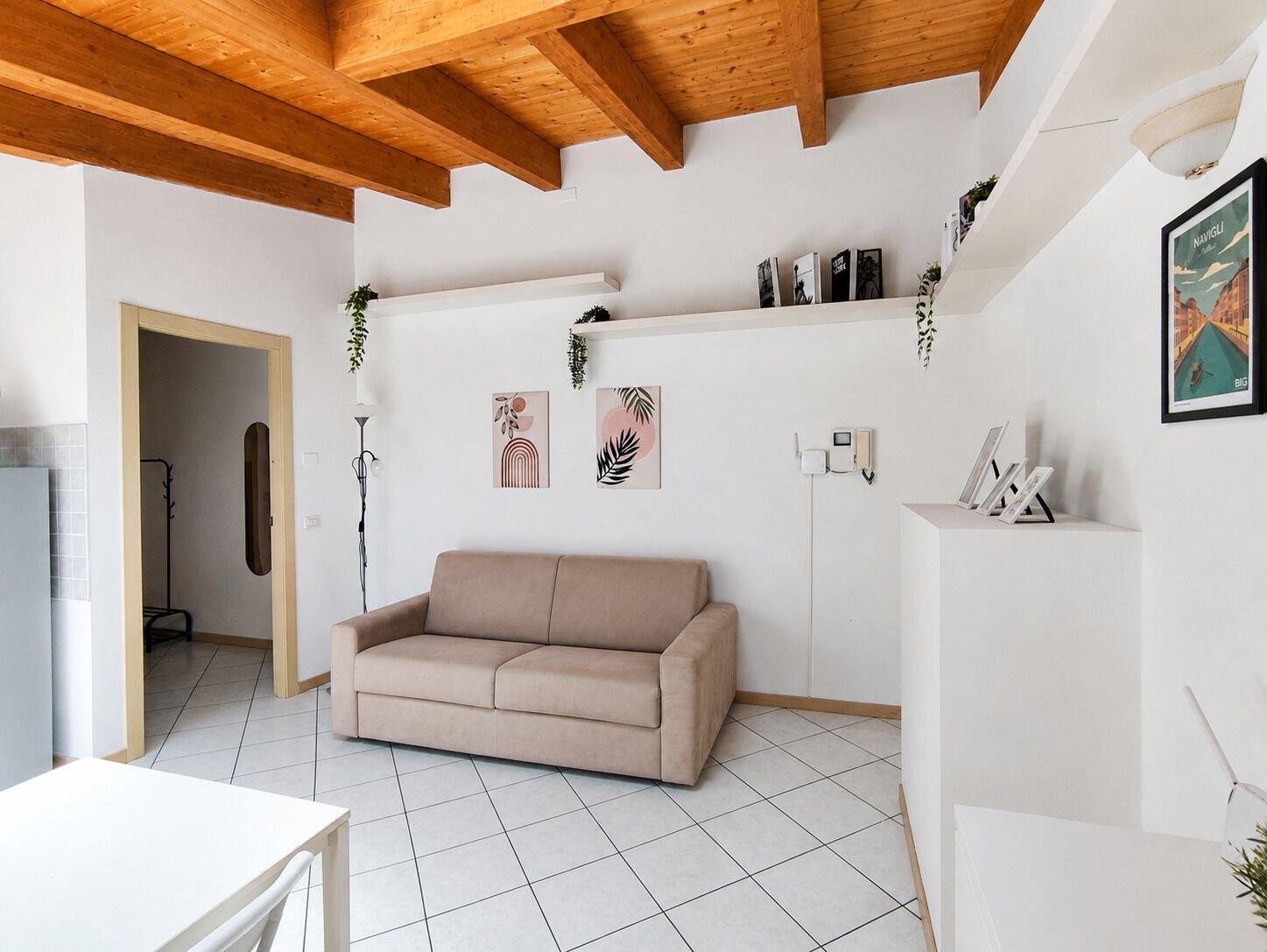 Un soggiorno con un divano color beige, pavimento in piastrelle bianche, travi a vista in legno e mensole a parete con piante.