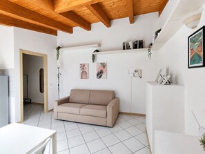 A living room with a beige sofa, white tiled floor, wooden ceiling beams, and wall-mounted shelves with plants.